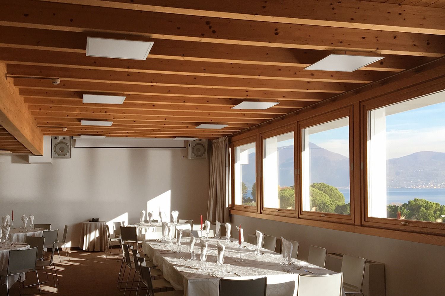 Camera con travi in ​​legno sul soffitto, ampie finestre con vista sul lago e sulle montagne e tavoli apparecchiati per i pasti.