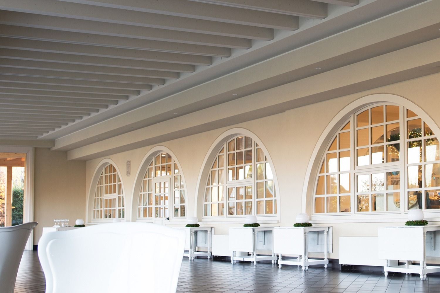 Uno spazio interno con finestre ad arco, pareti bianche e pavimento piastrellato. Tavoli e sedie bianchi sono visibili.