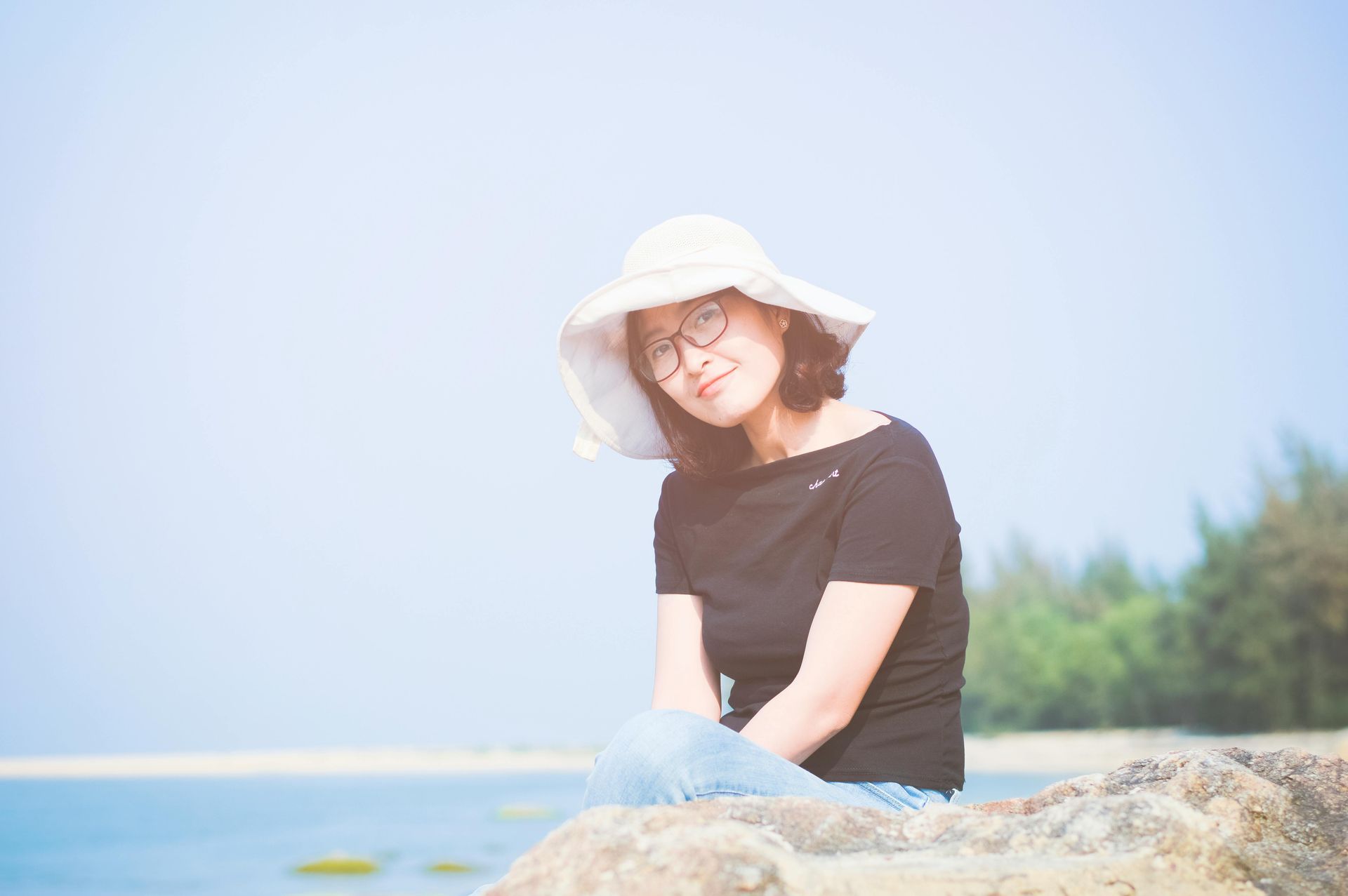 A woman wearing a hat and glasses is sitting on a rock near the ocean.