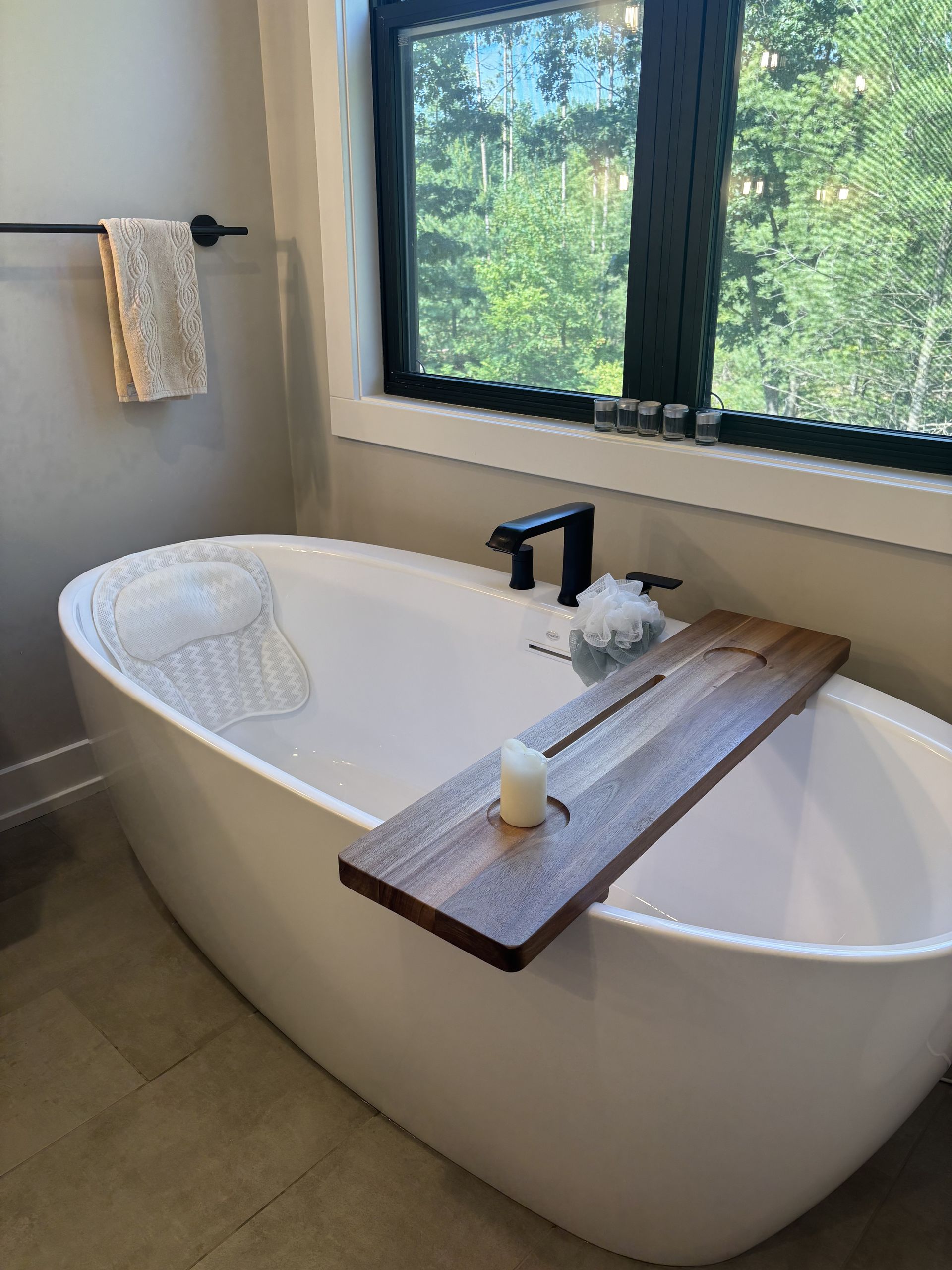 Oval white bathtub with a wooden tray, candle, and washcloth by a window.
