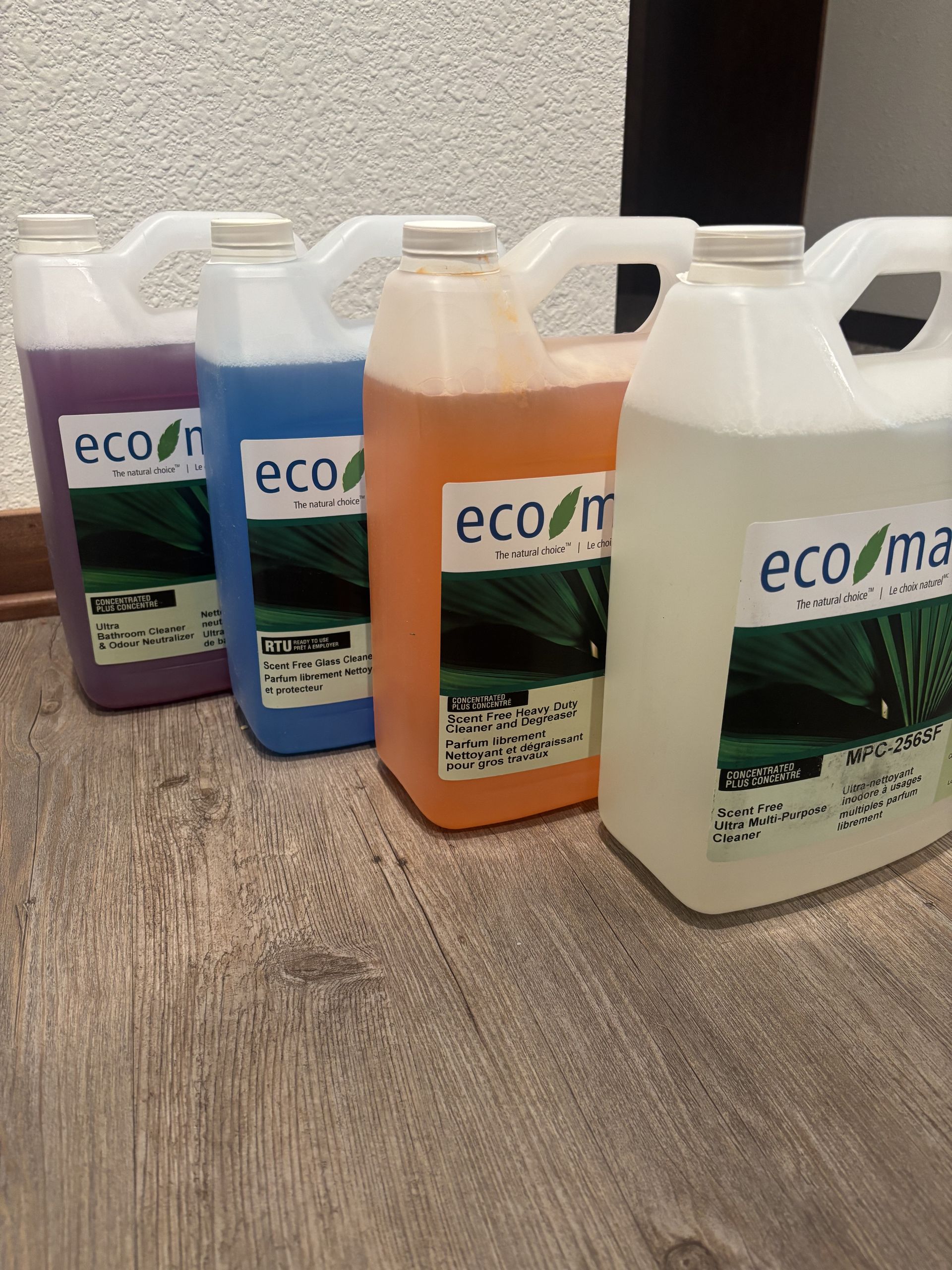 Four colorful bottles of eco-friendly cleaning products: purple, blue, orange, and clear, lined up on a wood floor.