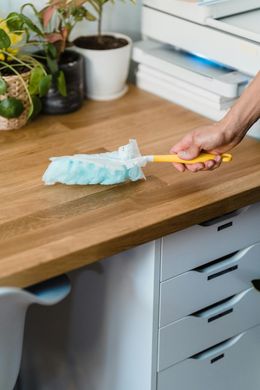 Person dusting a wooden desk with a blue and yellow duster. White drawers and plants are in the background.