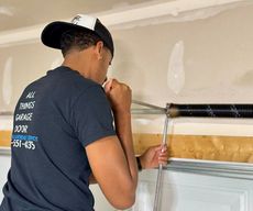 Man in black shirt and hat adjusting garage door spring. White garage interior.