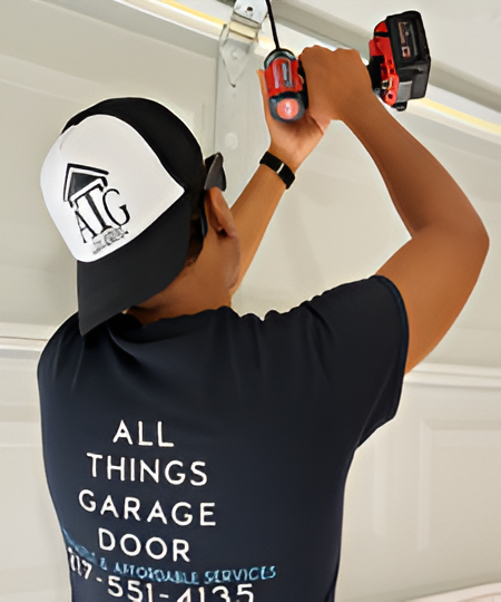 Person in black shirt and hat with ATG logo, using a drill to work on a garage door.