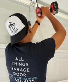 Man in a cap and t-shirt, working on a garage door with a drill.