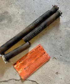 Three rusty garage door springs and two orange warning packets on concrete.
