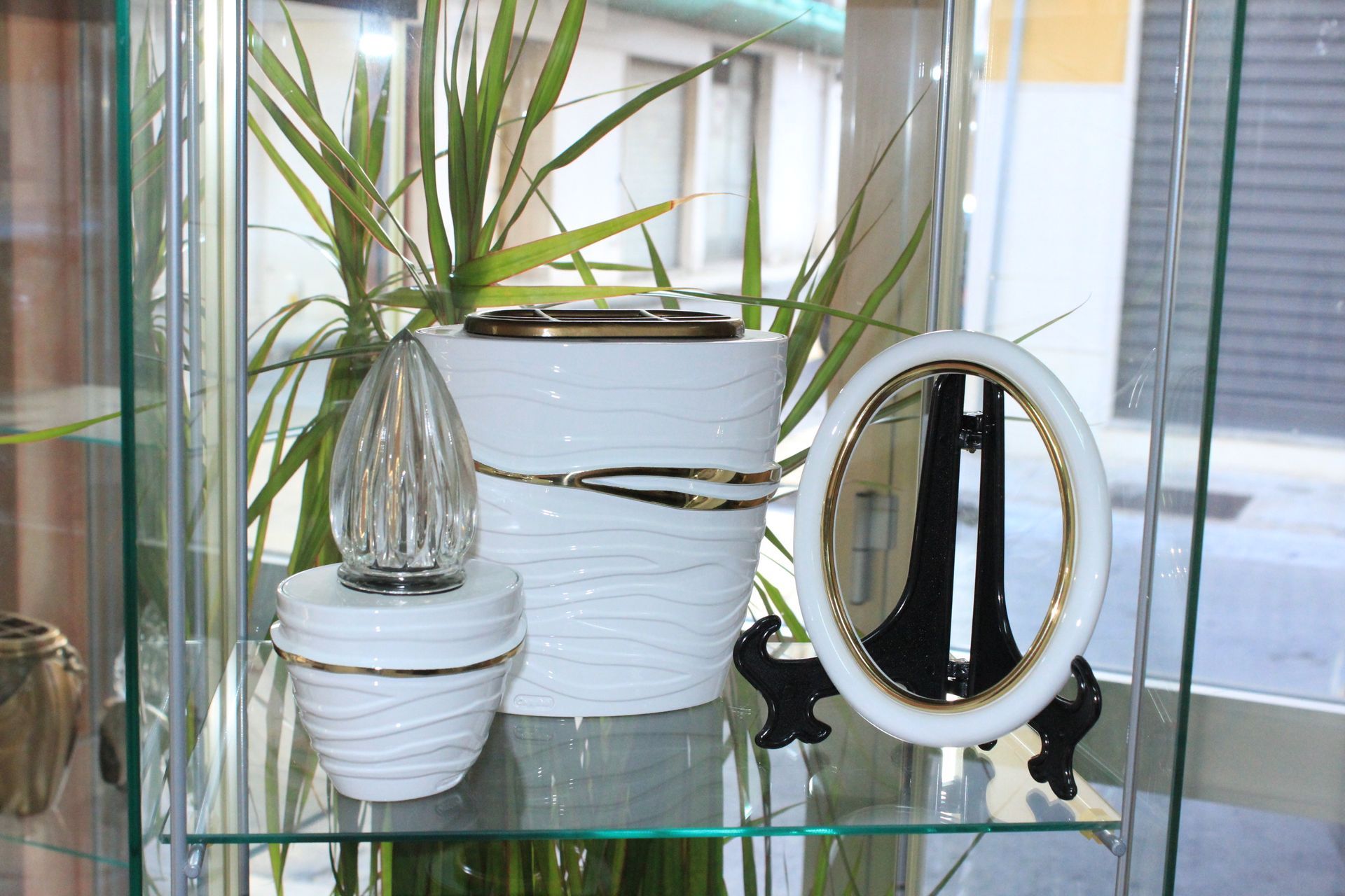 Urne bianche e dorate e una cornice su una mensola di vetro con una pianta sullo sfondo.
