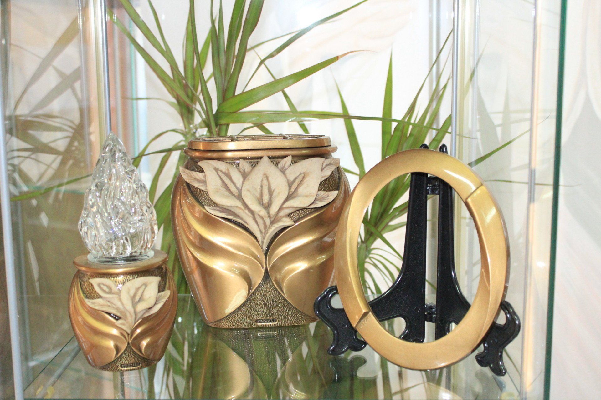 Vasi color oro e crema con una pianta sullo sfondo e una cornice ovale su un supporto in una teca di vetro.