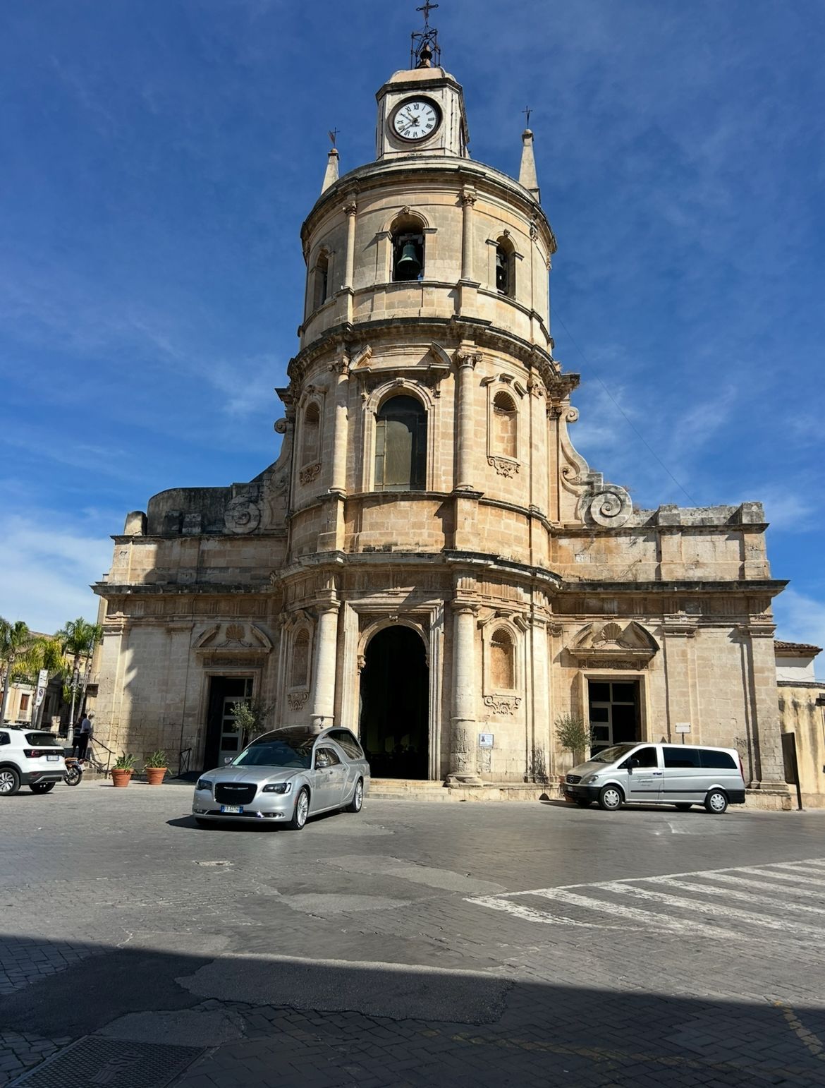 Tre uomini in abito nero sono in piedi accanto a un carro funebre argentato di fronte a un edificio con gradini.