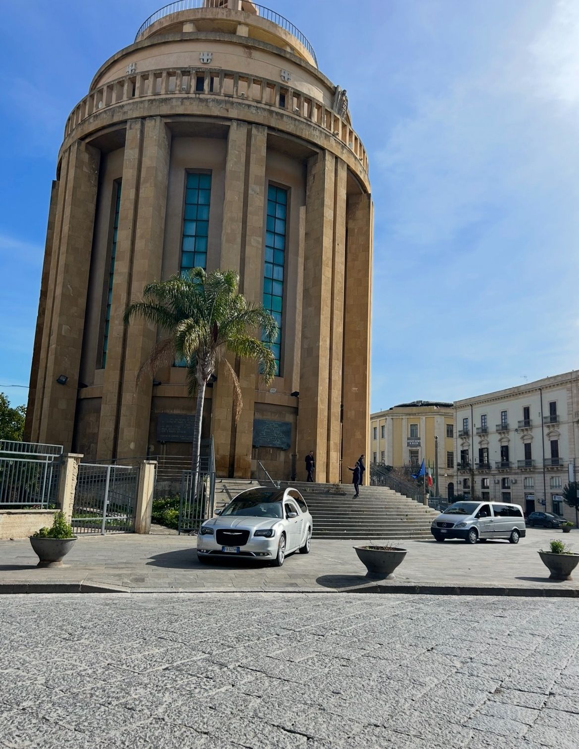 Tre uomini in giacca e cravatta sono in piedi davanti a carri funebri e a una chiesa in una giornata di sole.