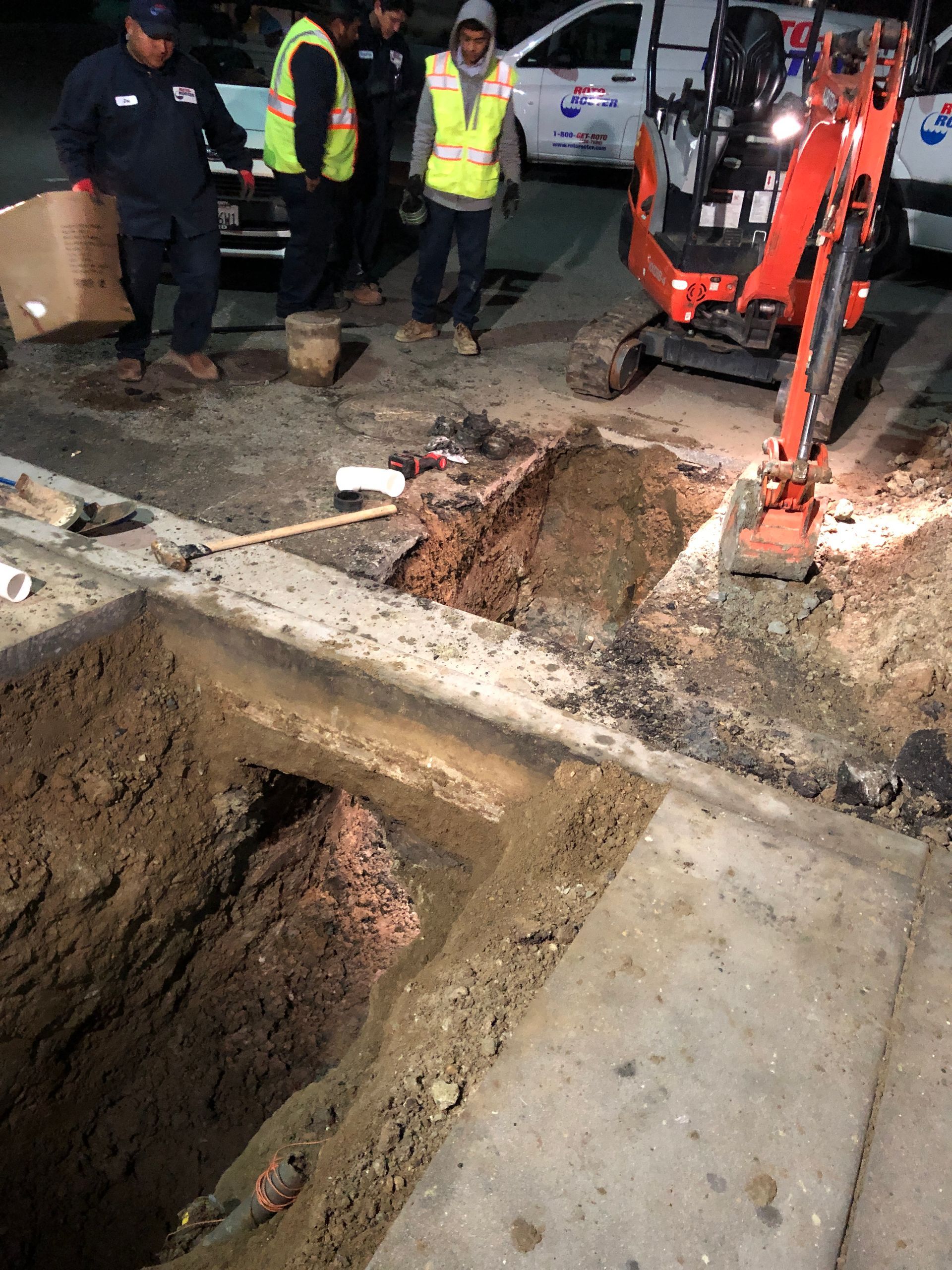 Workers around an excavation site, with a mini excavator, at night.