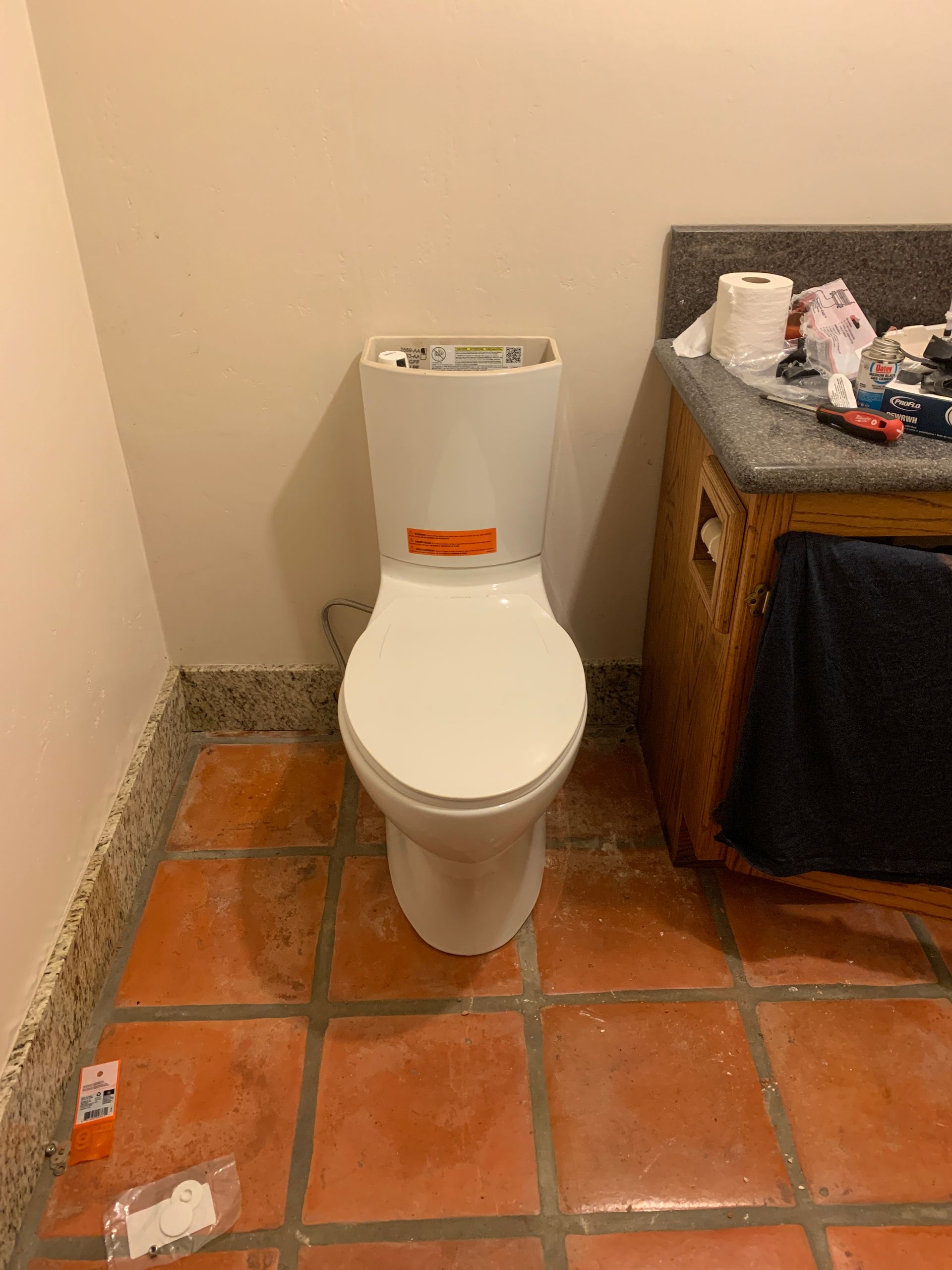 White toilet installed against a light-colored wall, on terracotta tile floor. Tools on countertop nearby.