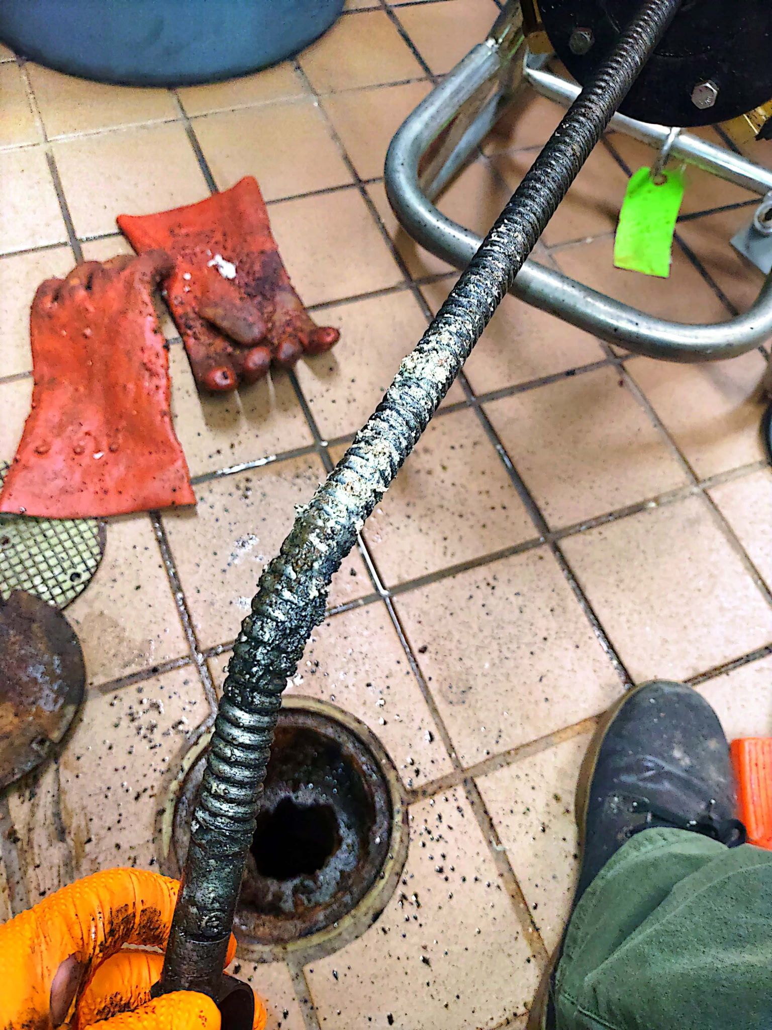 Plumber's snake covered in debris, held above an open drain on tiled floor, orange-gloved hand, red gloves.