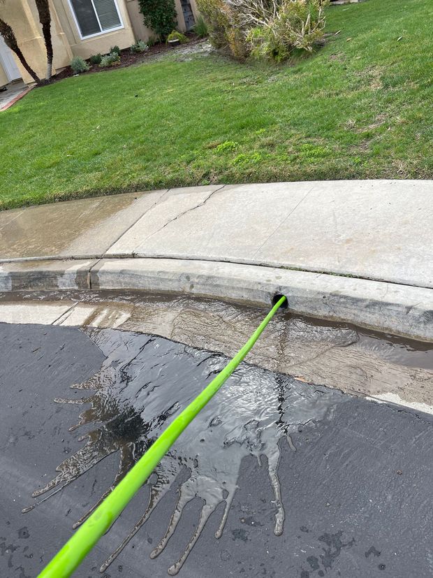 Man spraying water into a sewer drain with a pressure washer on a brick-lined street.