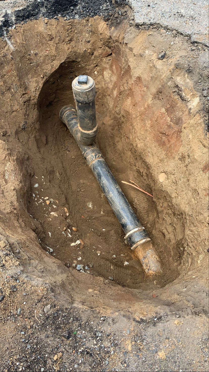 Trench in asphalt reveals black pipe with a T-joint and a copper wire.