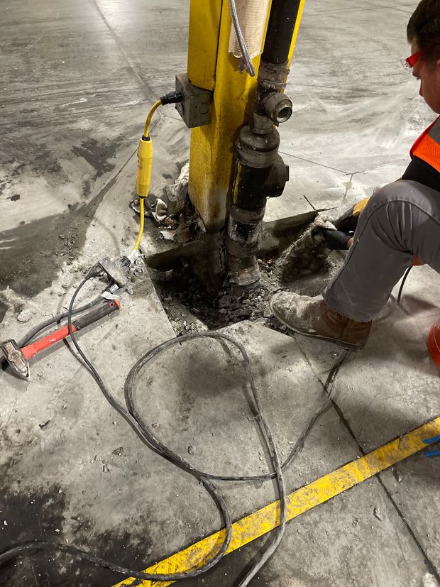 Worker in a yellow hard hat inspects a pipeline in a trench outdoors. A tractor is behind him.
