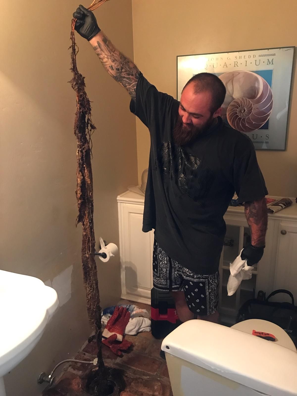 Man in bathroom holds long clog pulled from toilet. Dark hair, tattoos, smiling. White cabinets and sink.