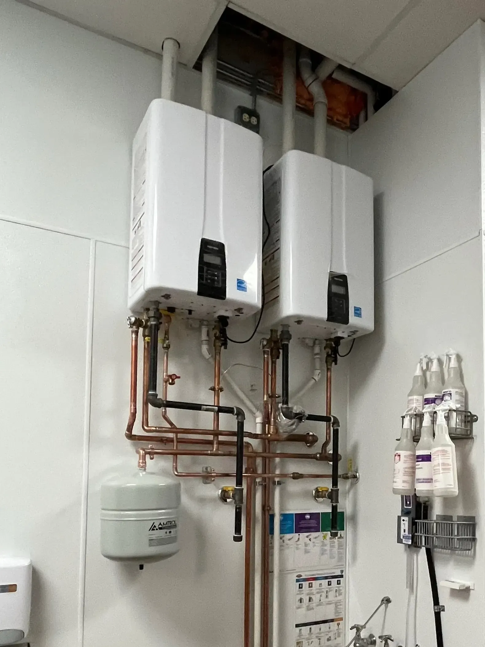 Two white tankless water heaters with copper pipes mounted on a white wall.