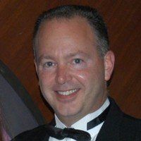 Man in black tuxedo smiling, dark hair, against a brown background.