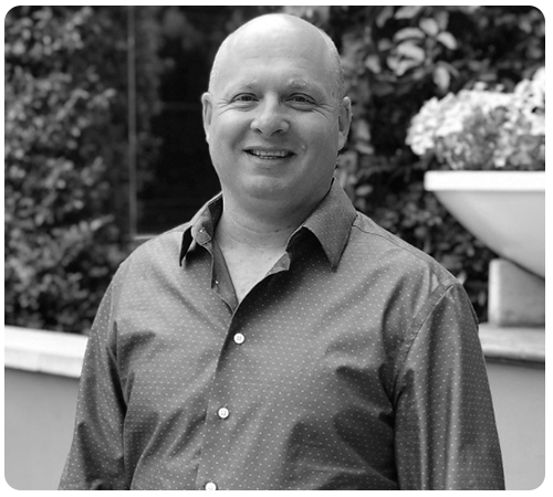 Man in a patterned button-down shirt smiles, standing outdoors near foliage.