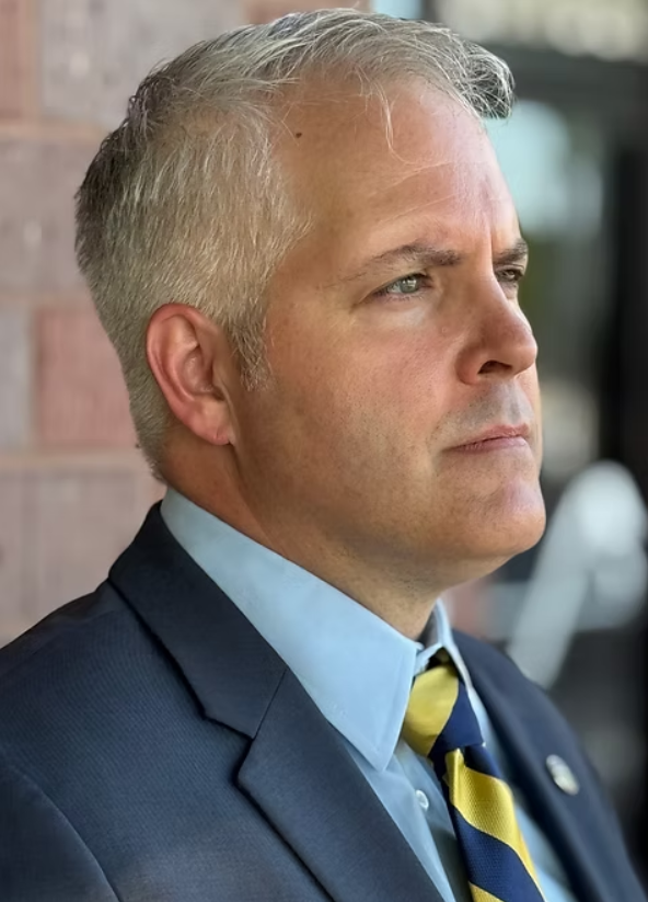Man in a blue suit with yellow and blue tie, looking off-camera with a serious expression.