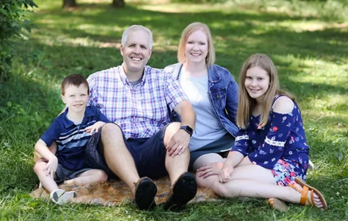 Family of four smiles while seated on a rug in grassy area.