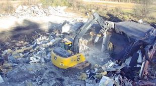 A yellow excavator is demolishing a building in a field.