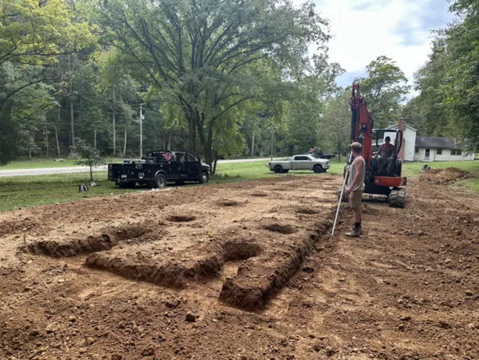 A man is standing in the middle of a dirt field next to an excavator.