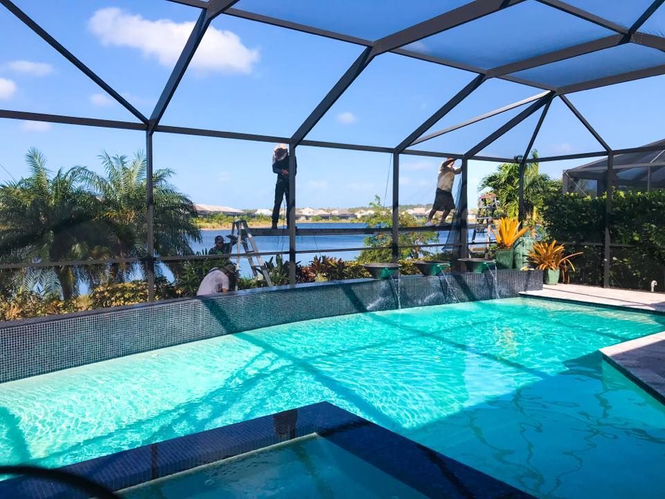 Workers repairing a screened-in pool enclosure overlooking a pool and a lake under a bright blue sky.