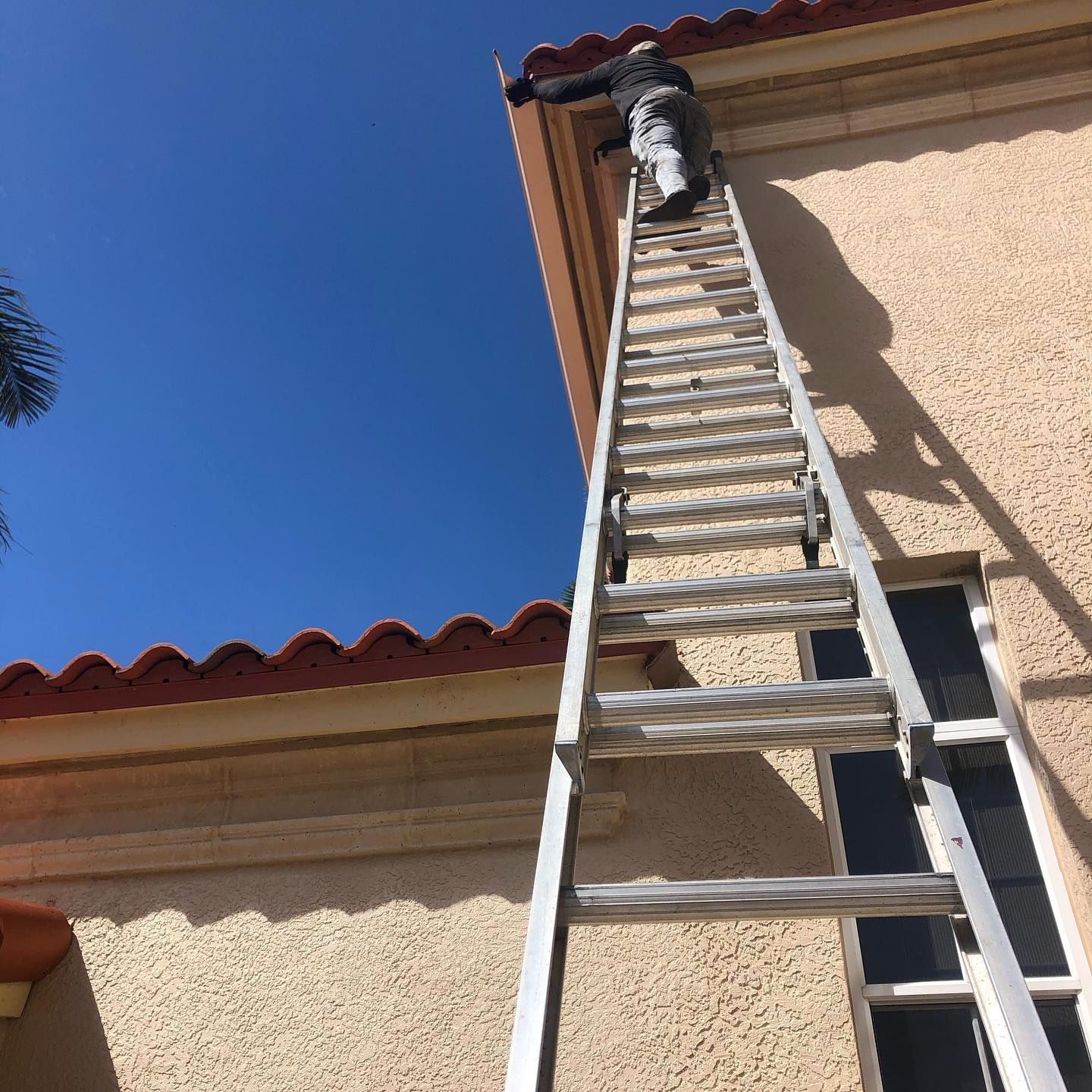 Person on a tall ladder painting the edge of a tan stucco building with a red tile roof against a blue sky.