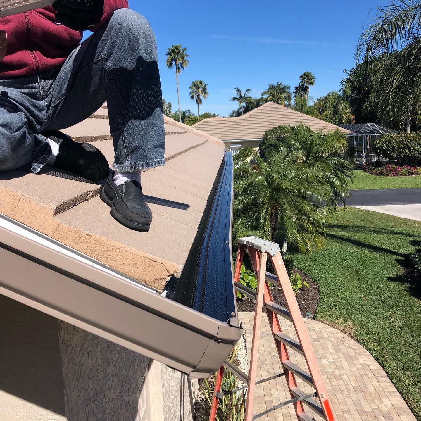 Person on a roof installing a gutter; orange ladder nearby; sunny day with palm trees.