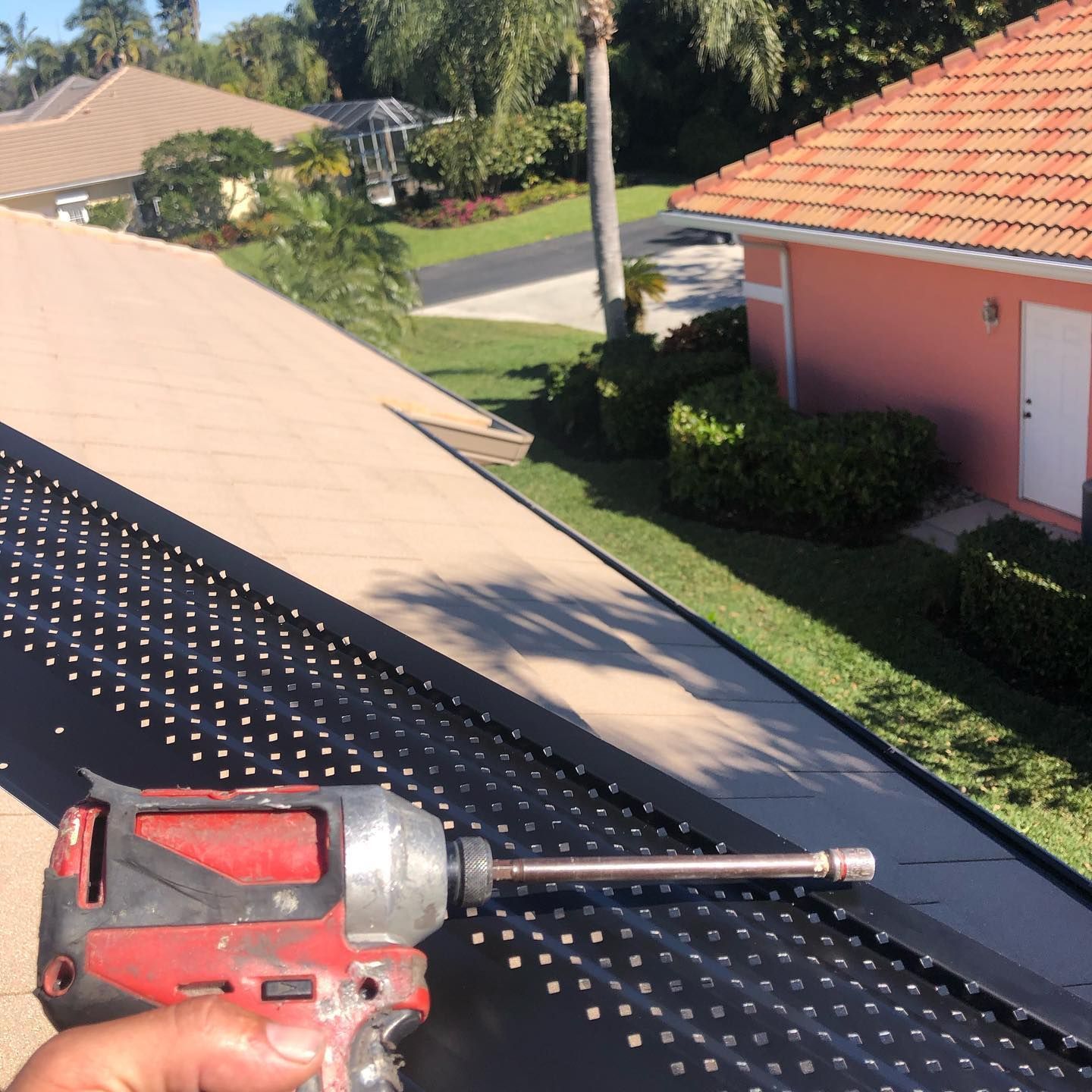 Hand holding a red power drill installing black gutter guards on a roof with a view of a neighborhood.