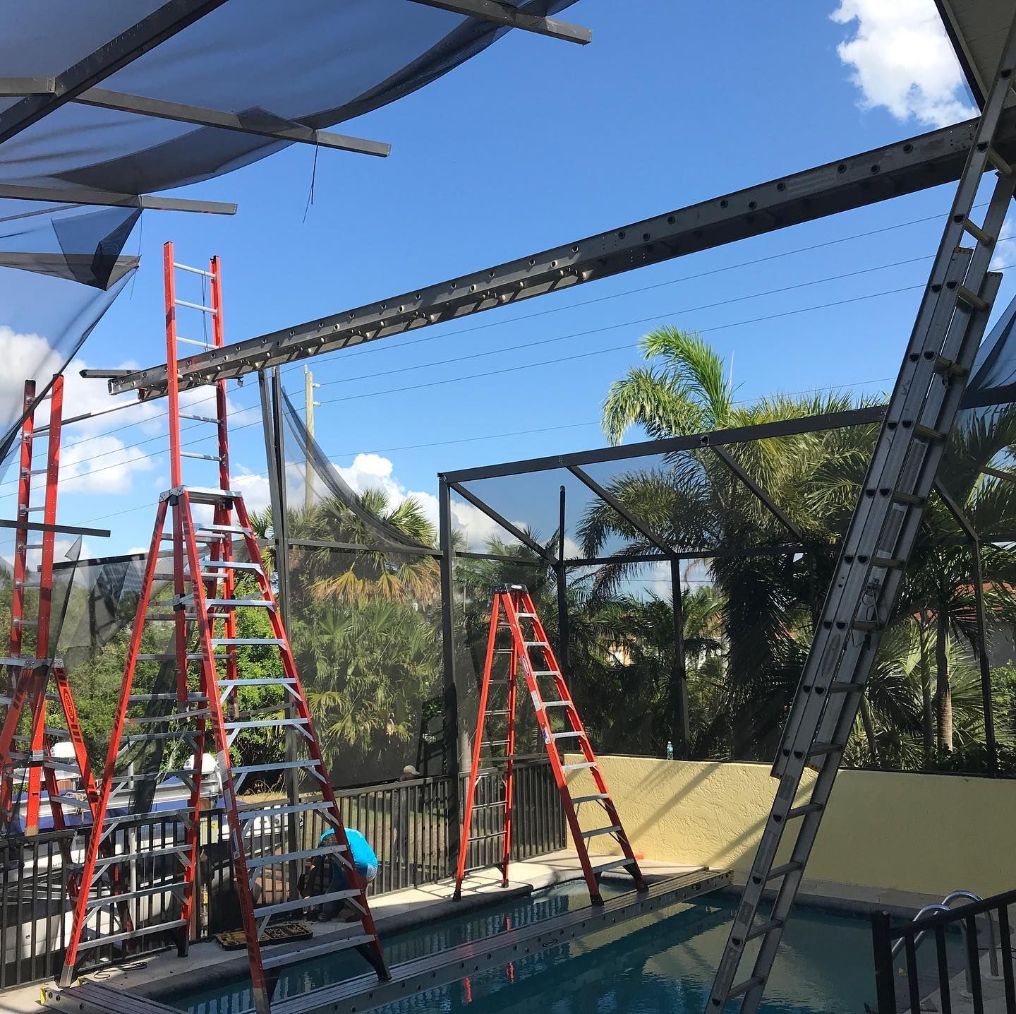 Pool enclosure construction: ladders, black metal framing, blue pool, sunny day.
