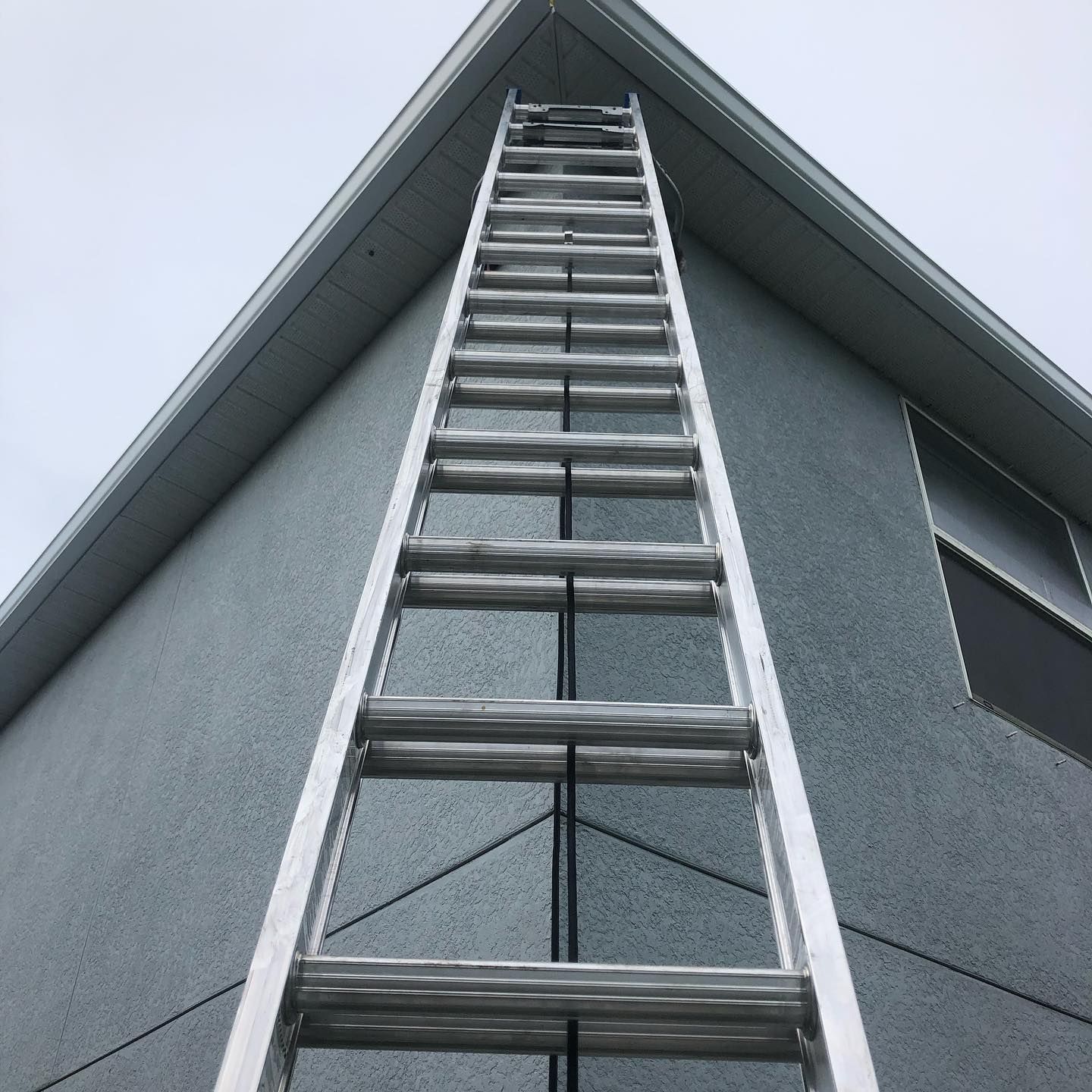 Ladder leaning against a gray building with dark window and light sky.