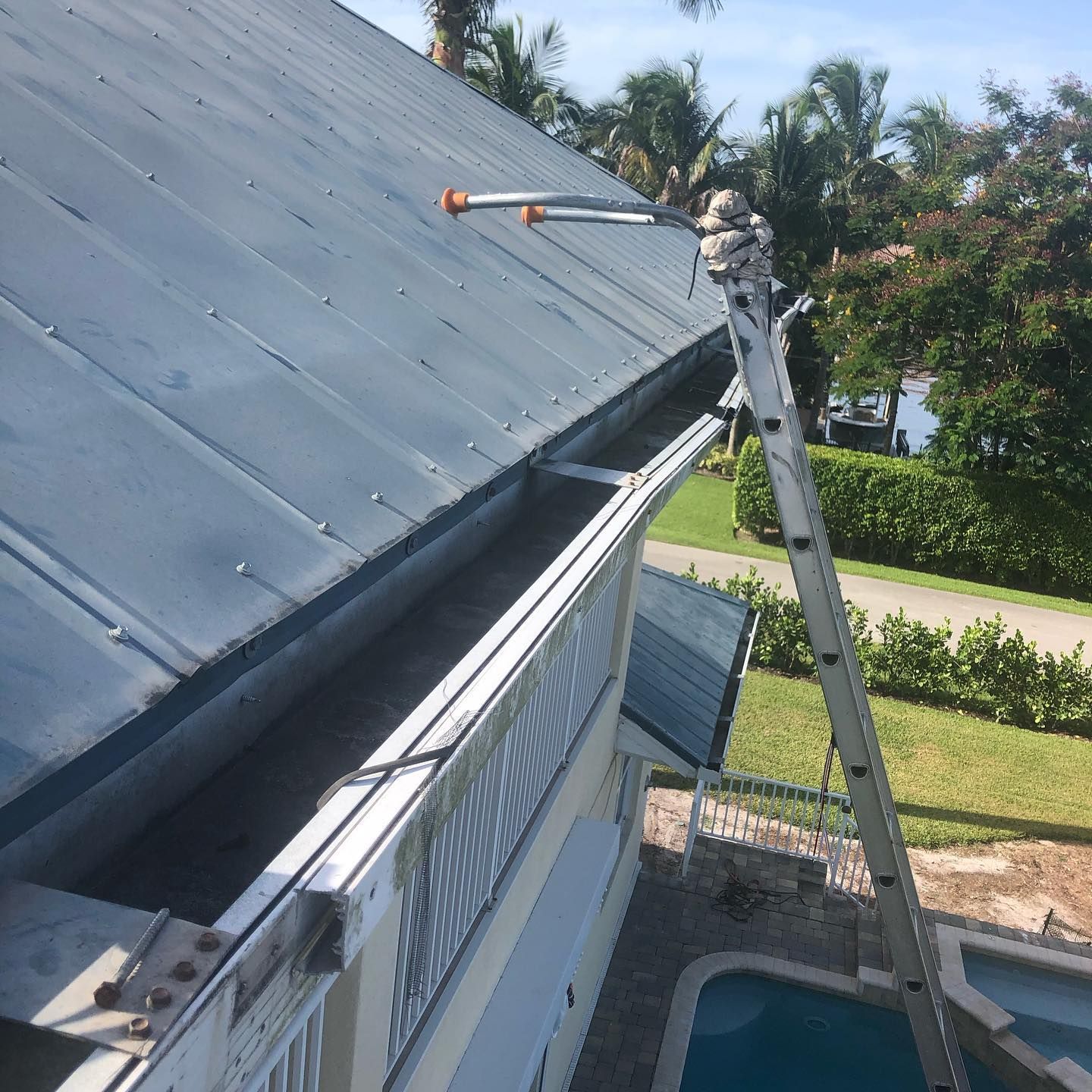 Ladder on roof next to a gutter; cleaning, exterior view. Blue roof, sunny day, pool below.