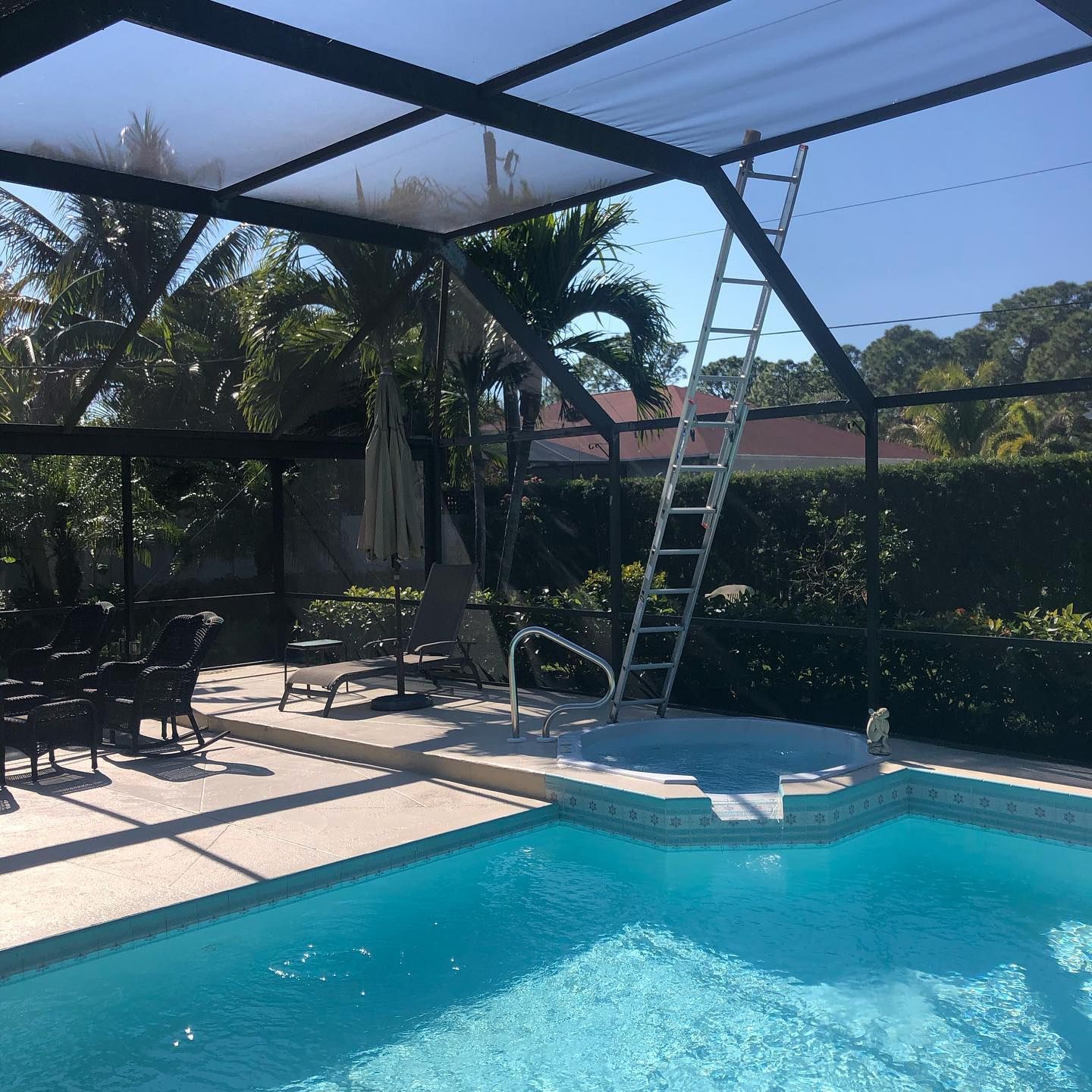 Pool area with ladder against screen enclosure, blue water, outdoor furniture, and trees.