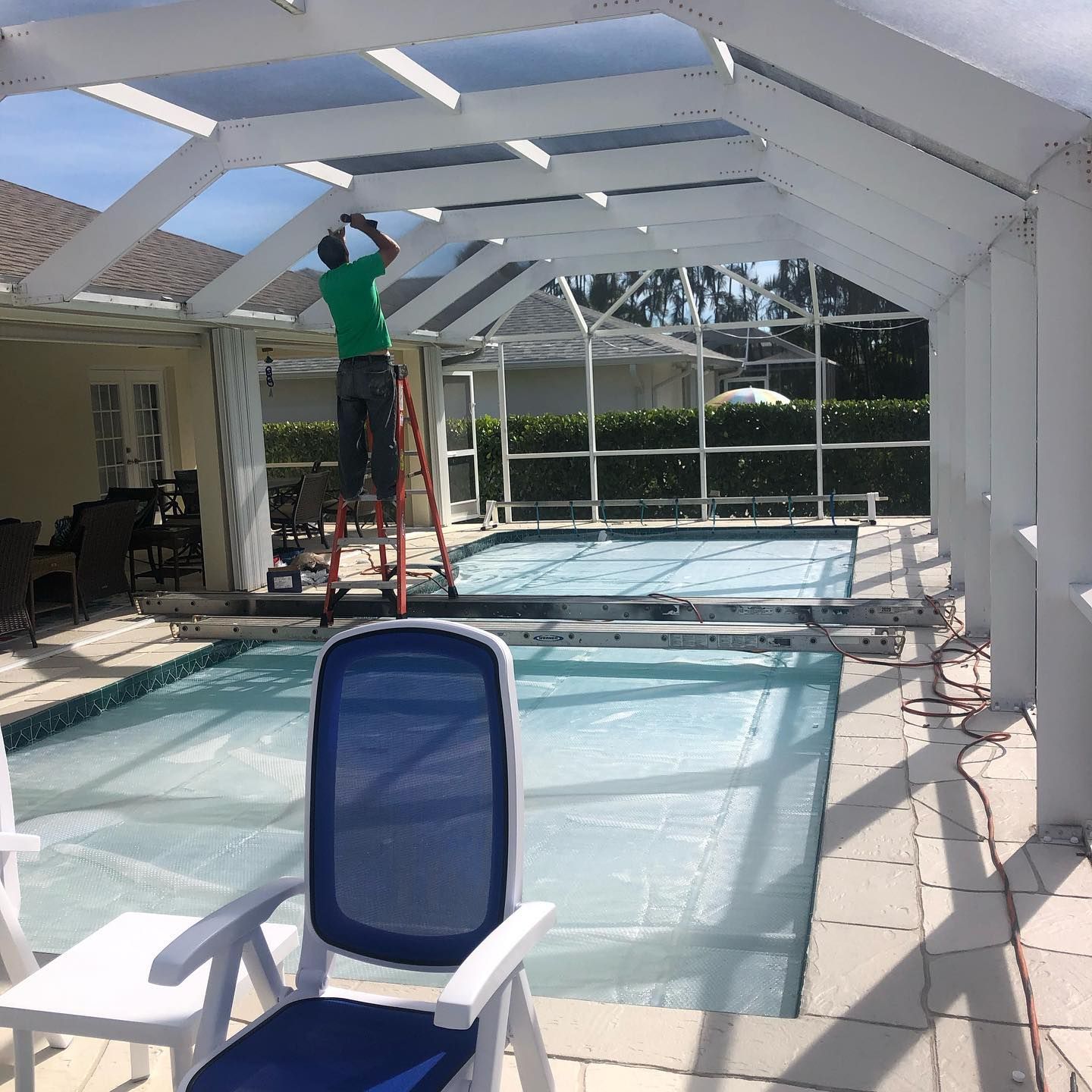 A person on a ladder working on a white-framed pool enclosure. The pool is covered with a blue tarp.