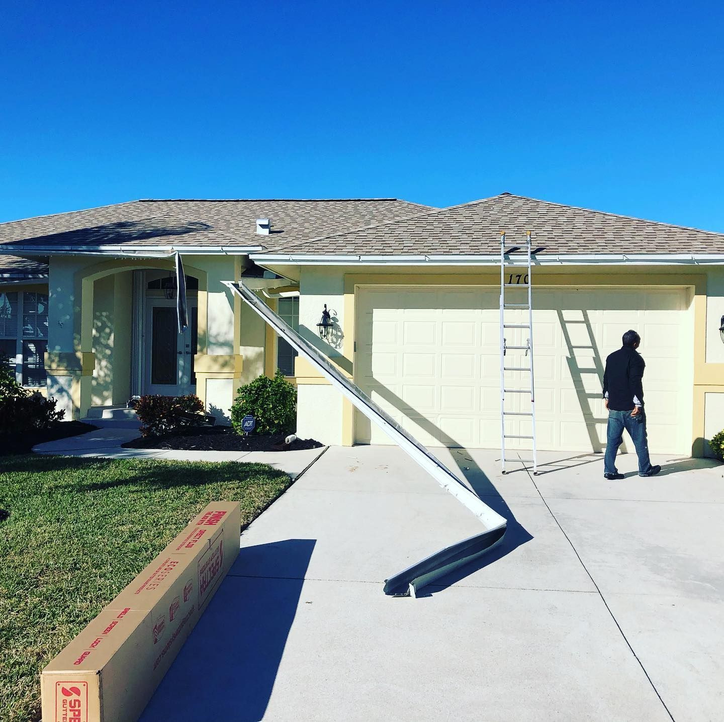 A person installing a gutter near a home; ladder and gutter material are present on the driveway.