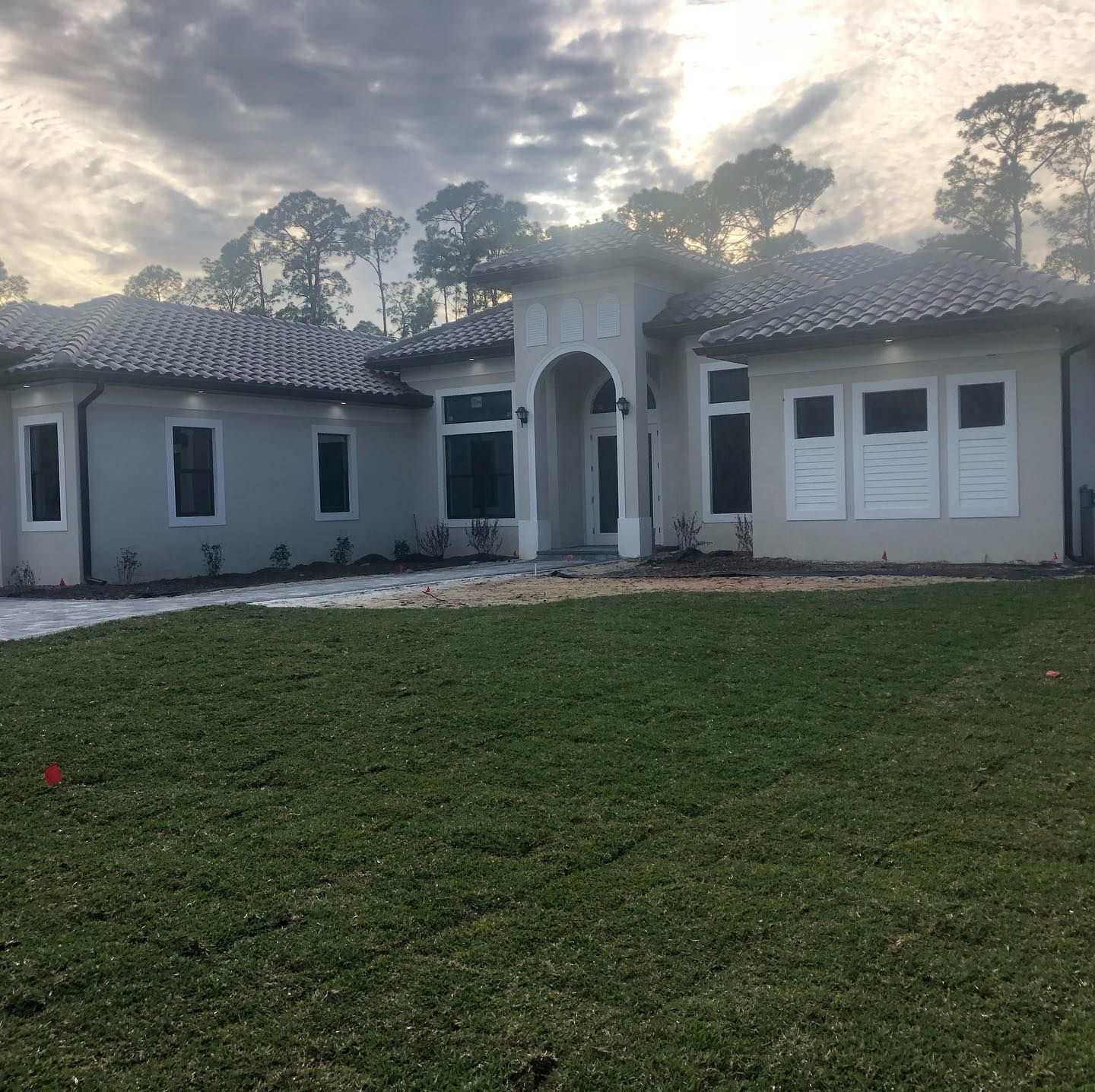 A light gray stucco house with a tile roof, arched entryway, and dark-framed windows against a cloudy sky.