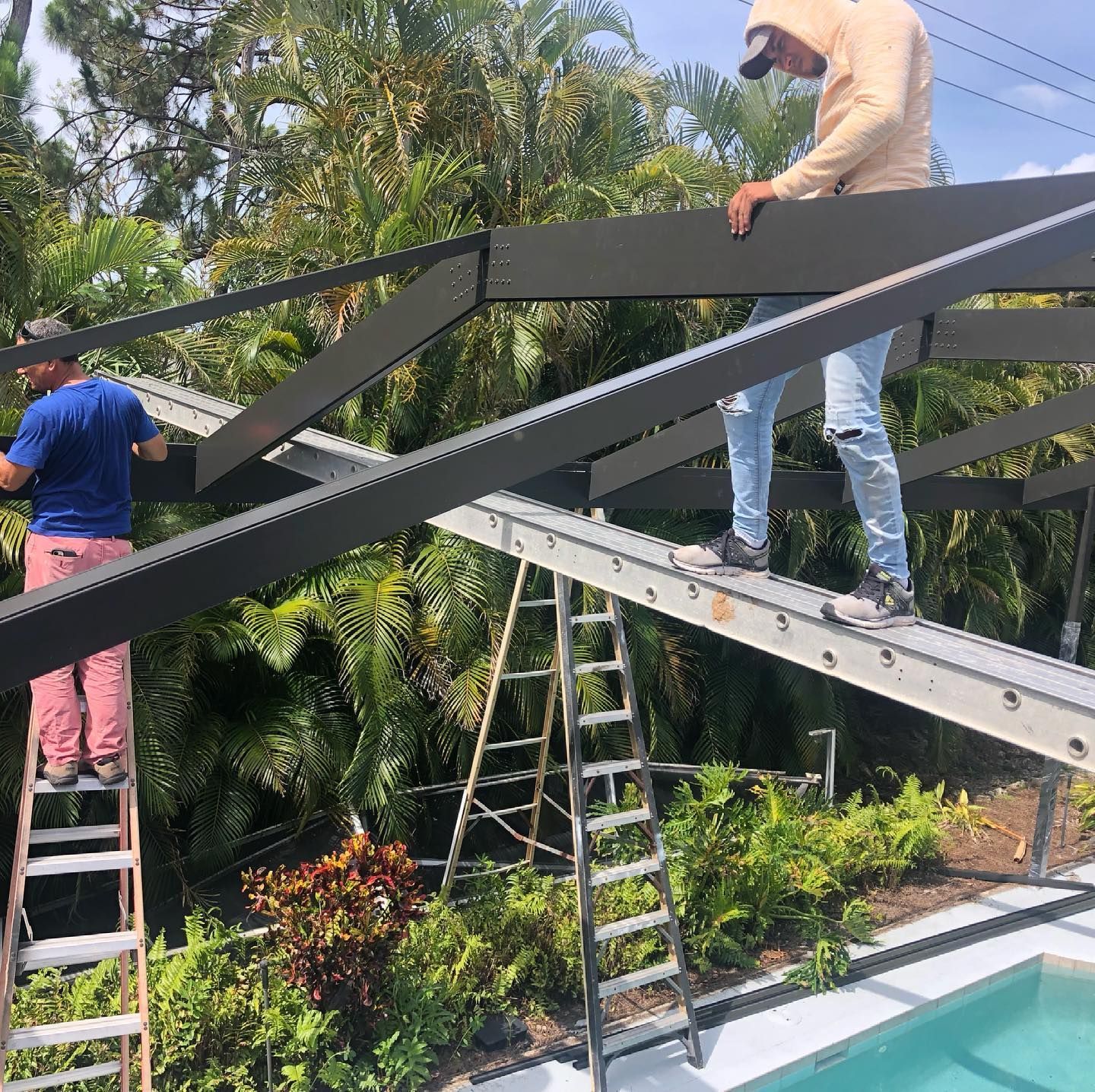 Two workers constructing a black-beamed structure over a pool, using ladders. Sunny outdoor setting with lush plants.