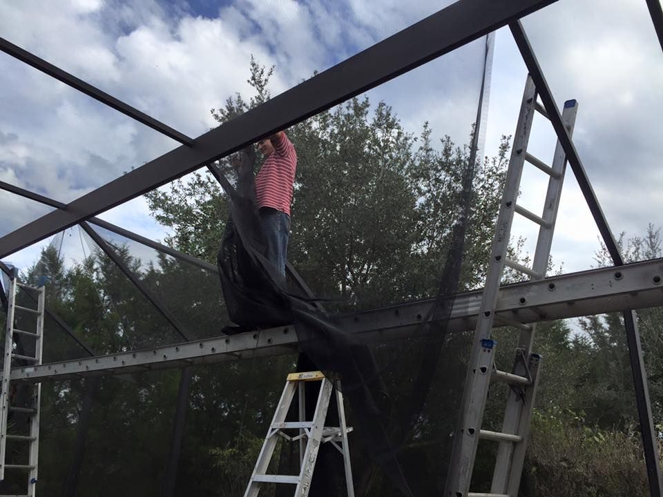 Person on a ladder installing black screening on a metal frame, against a cloudy sky.