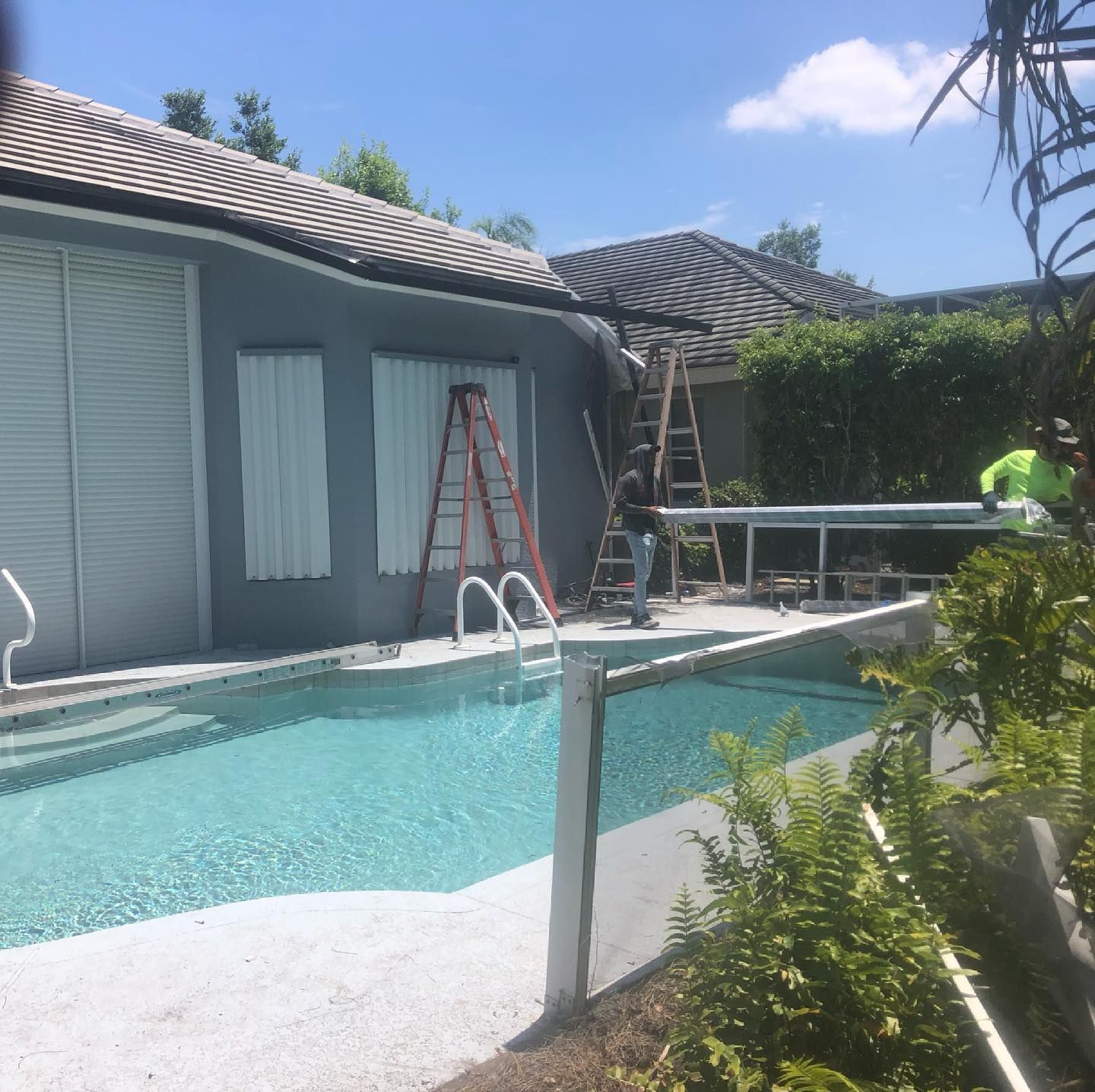 Workers near a pool, repairing house siding. Blue water, gray house, ladders. Sunny day.