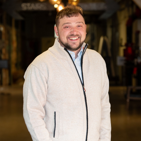 Smiling man in a tan zip-up sweater and blue shirt, in an industrial setting with a