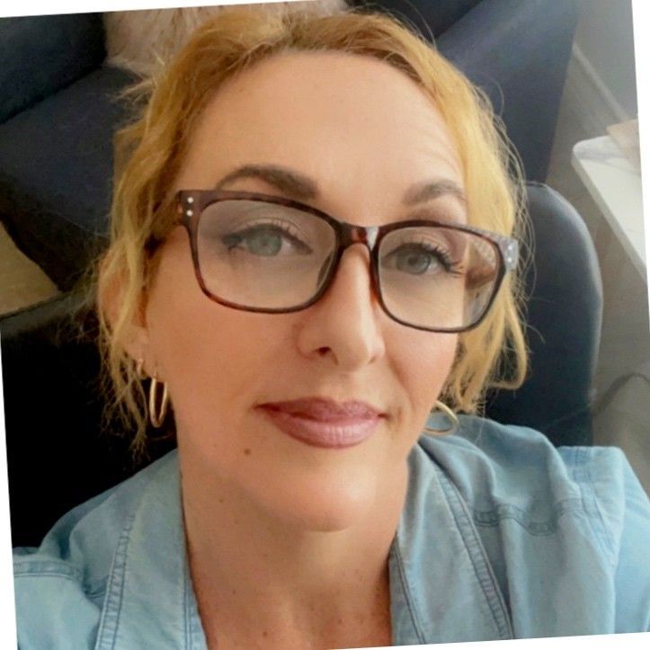 Woman with blonde hair and glasses smiling. Wearing a blue shirt, indoors.