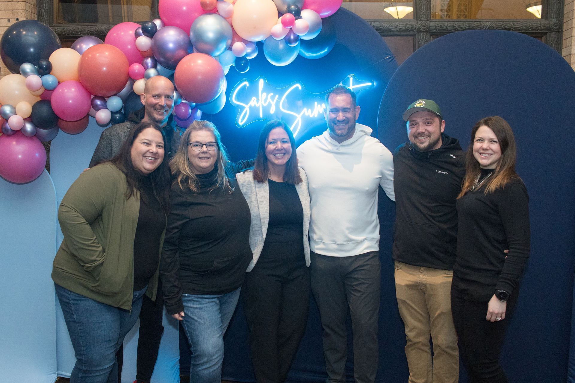 Five smiling people standing in front of a white brick wall; three women and two men, wearing jeans and shirts.