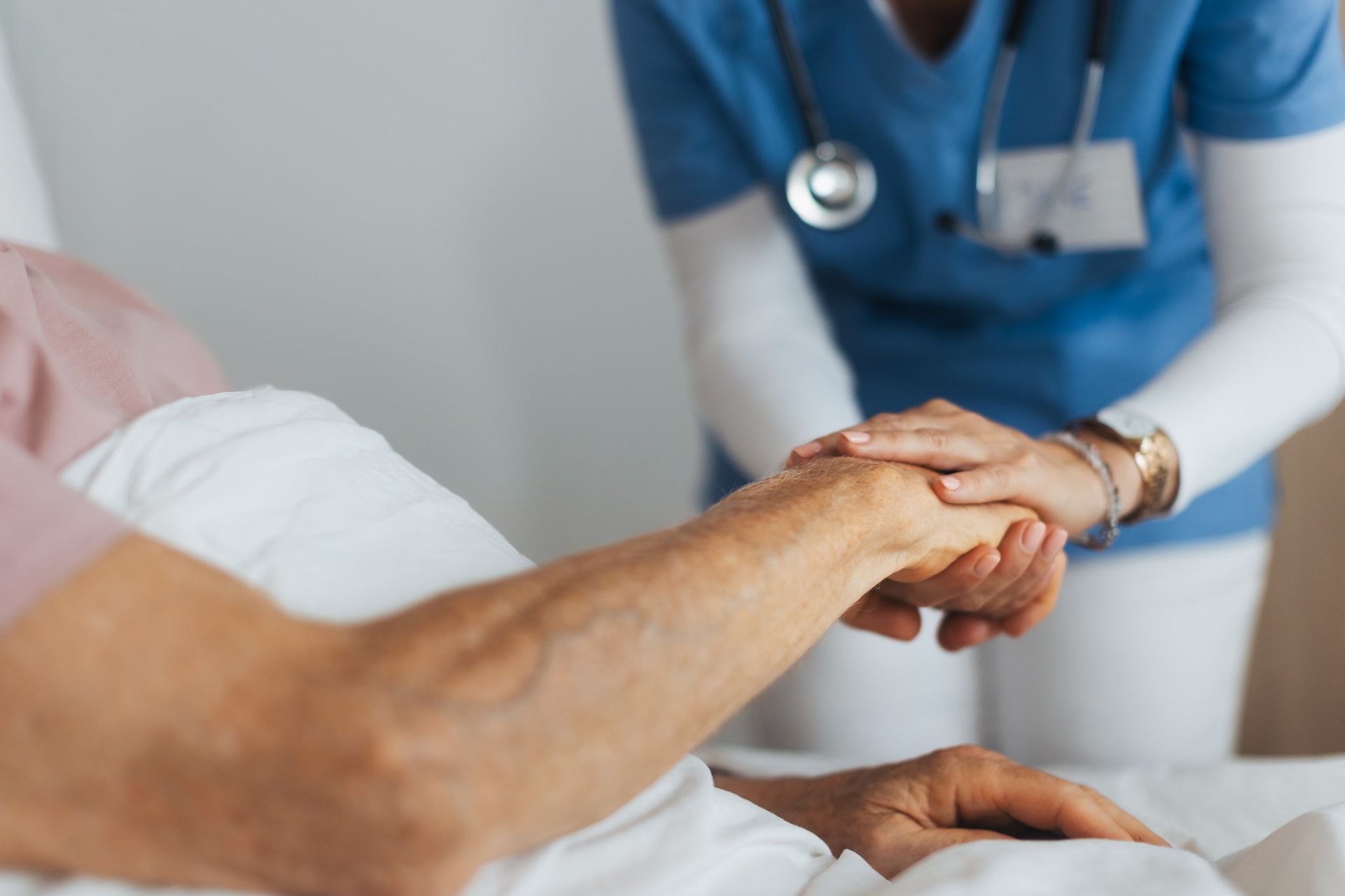 A nurse in a blue uniform holds the hand of an elderly man, showcasing palliative care services.