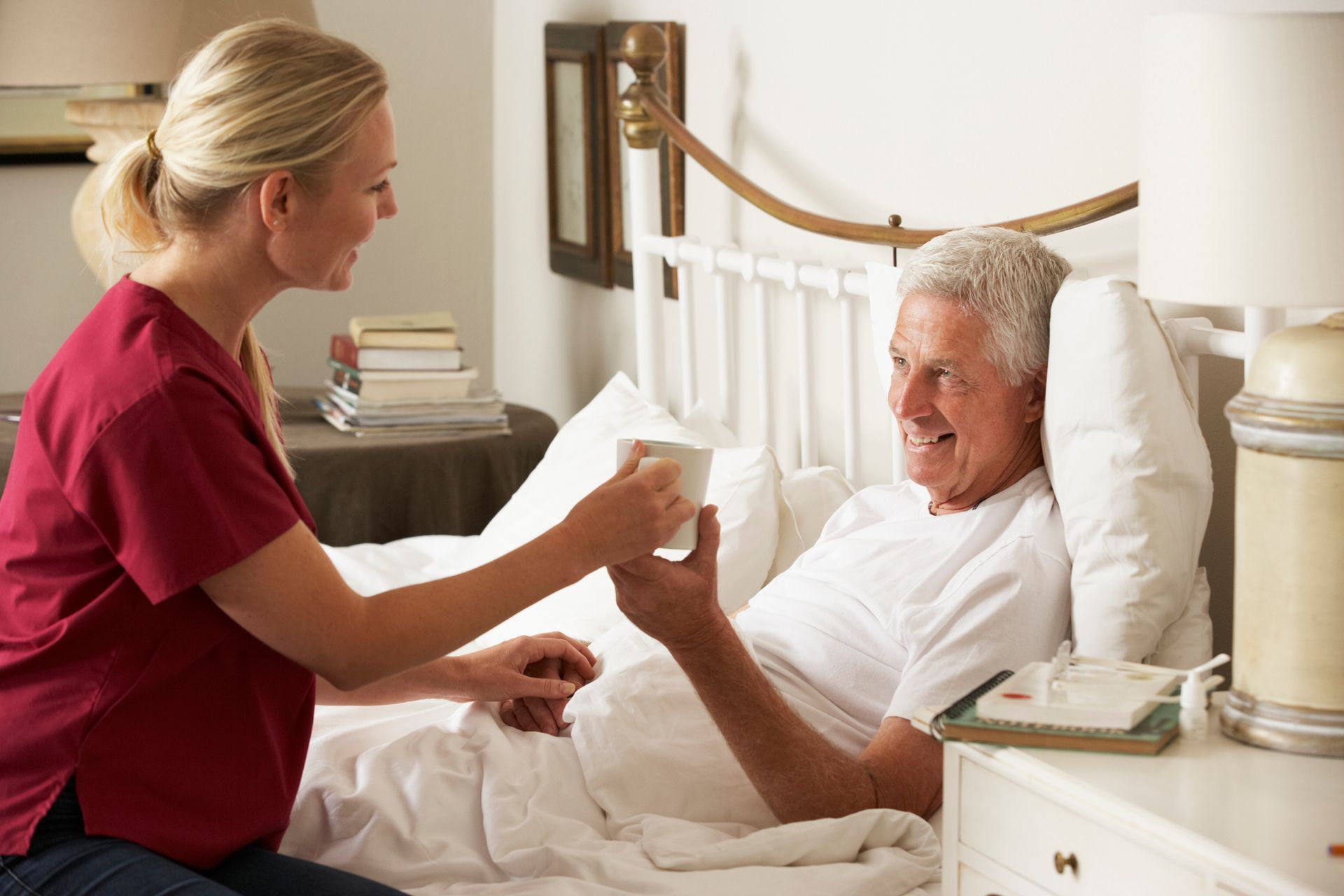A female nurse in red scrubs hands a male elder a drink in a cup as he smiles and lays in a bed.