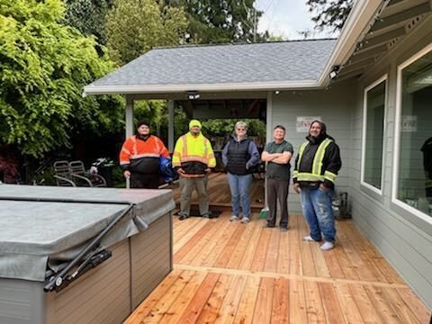 Five people standing on a wooden deck next to a hot tub. Some wear work vests. Grey house, green trees.