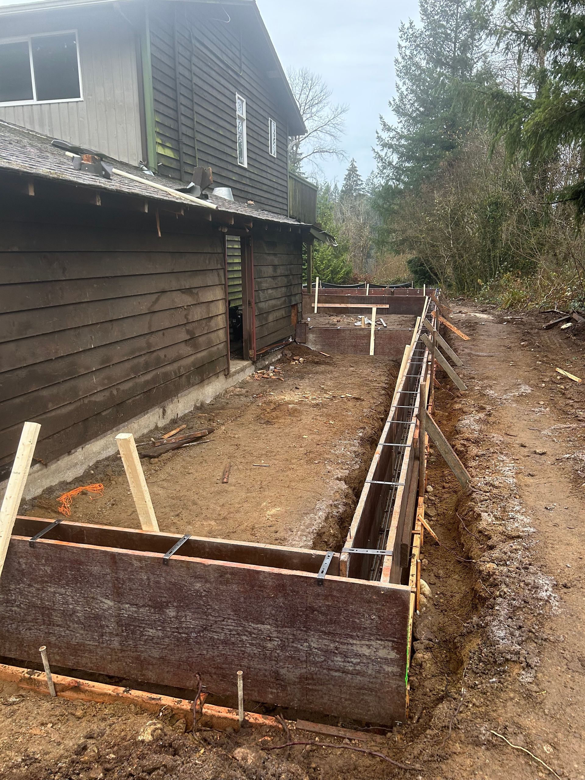 Wooden forms for a concrete foundation being constructed next to a two-story building in an outdoor setting.