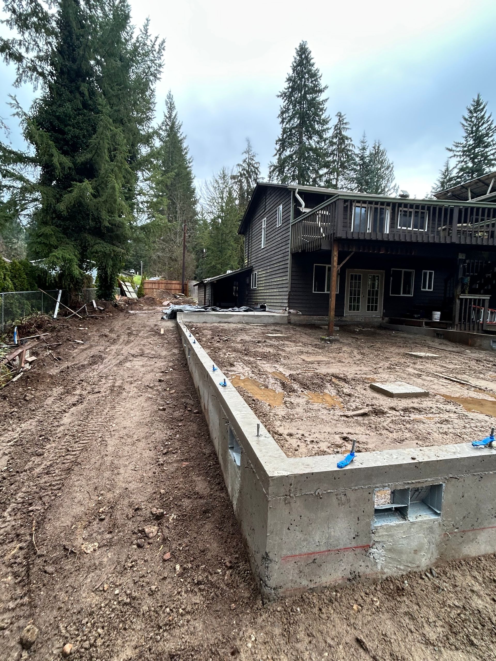 Concrete foundation with exposed rebar in muddy yard; house with burnt siding in background.